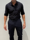 A man wearing a black long-sleeve button-down shirt with rolled-up sleeves and dark trousers, standing against a plain light gray wall.