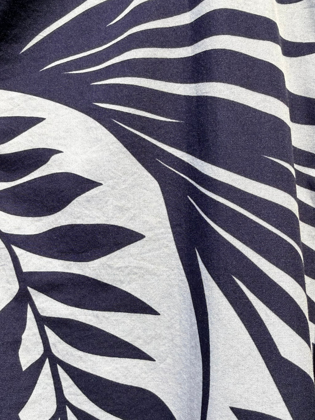 Close-up of a tropical leaf print fabric with dark navy blue palm fronds on a white background, showing part of a buttoned garment.