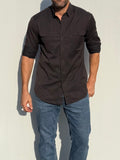 A man wearing a dark brown long-sleeve button-down shirt with chest flap pockets and blue jeans, standing against a light gray wall.