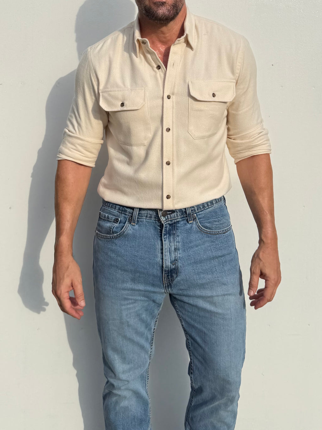 A man wearing a light beige long-sleeve button-up shirt with two chest pockets and light blue jeans, standing against a plain light wall.