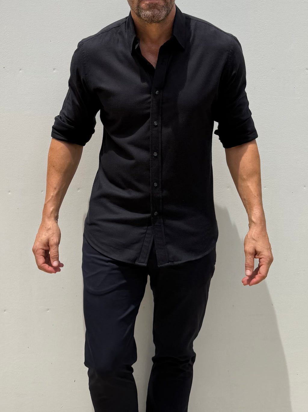 A man wearing a black long-sleeve button-down shirt with rolled-up sleeves and black pants, standing against a light gray wall.