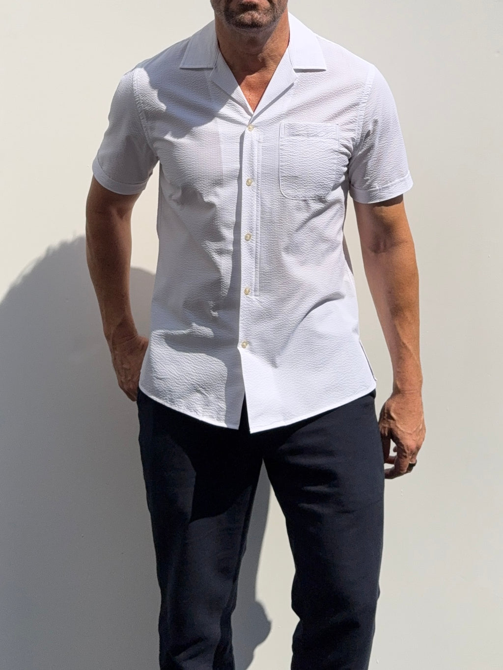 A man wearing a white short-sleeve button-up shirt with a subtle textured pattern and dark navy pants, standing against a light gray wall.
