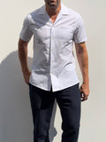 A man wearing a white short-sleeve button-up shirt with a subtle textured pattern and dark navy pants, standing against a light gray wall.