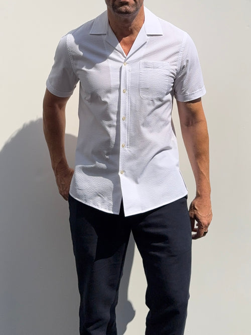 A man wearing a white short-sleeve button-up shirt with a subtle textured pattern and dark navy pants, standing against a light gray wall.