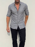 A man wearing a light gray long-sleeve button-down shirt with rolled-up sleeves and dark pants, standing against a plain off-white wall.