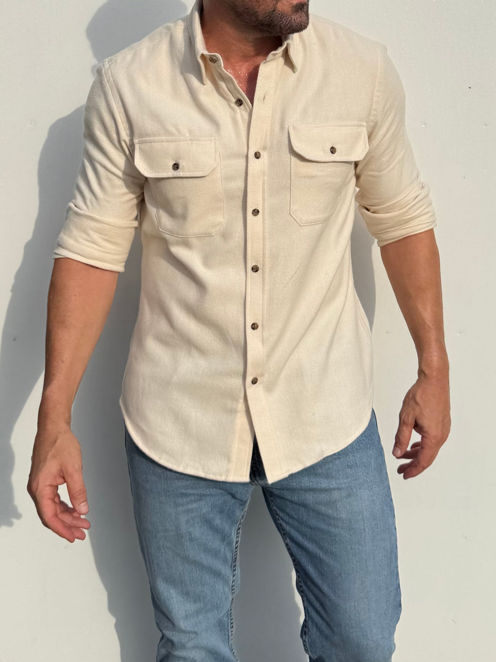 A man wearing a light beige linen button-up shirt with three-quarter sleeves and two chest pockets, paired with blue jeans, standing against a plain light gray wall.