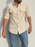 A man wearing a light beige linen button-up shirt with three-quarter sleeves and two chest pockets, paired with blue jeans, standing against a plain light gray wall.
