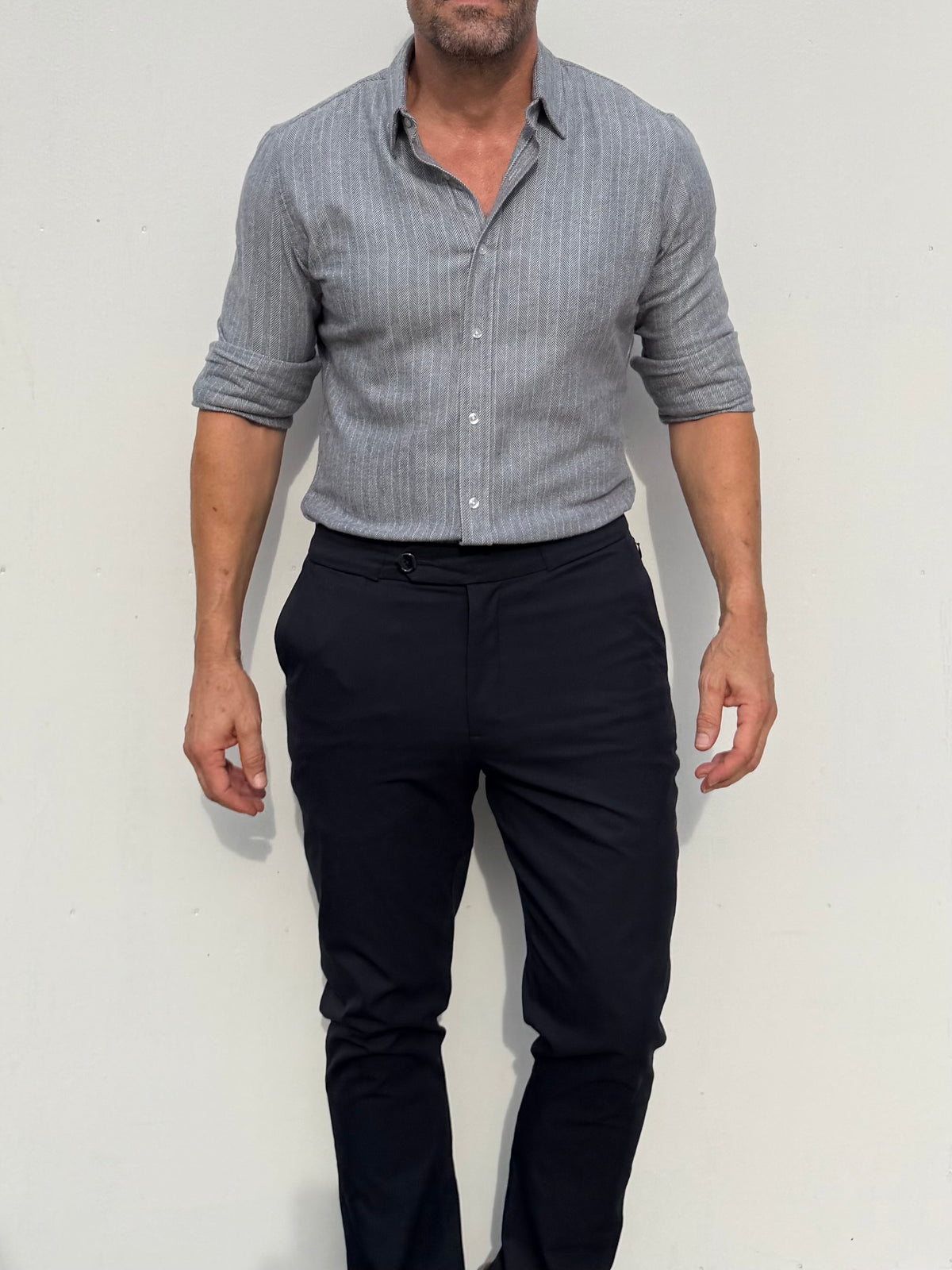A man wearing a light gray and white striped button-down shirt with rolled-up sleeves and dark navy trousers, standing against a plain off-white wall.
