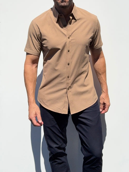 A man wearing a short-sleeve button-up shirt in a warm tan or camel color, paired with dark navy pants, standing against a plain white background.