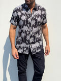 A man wearing a black short-sleeve button-up shirt with a white palm tree pattern, paired with black pants, standing against a light gray wall.