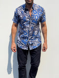 A man wearing a short-sleeve button-up shirt with a blue, white, and black paisley pattern, paired with dark pants, standing against a plain light background.